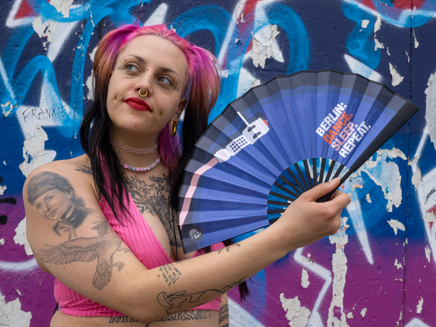 Person hält Fächer mit Design Berlin Dance Sleep Repeat in der Hand vor Graffiti Wand