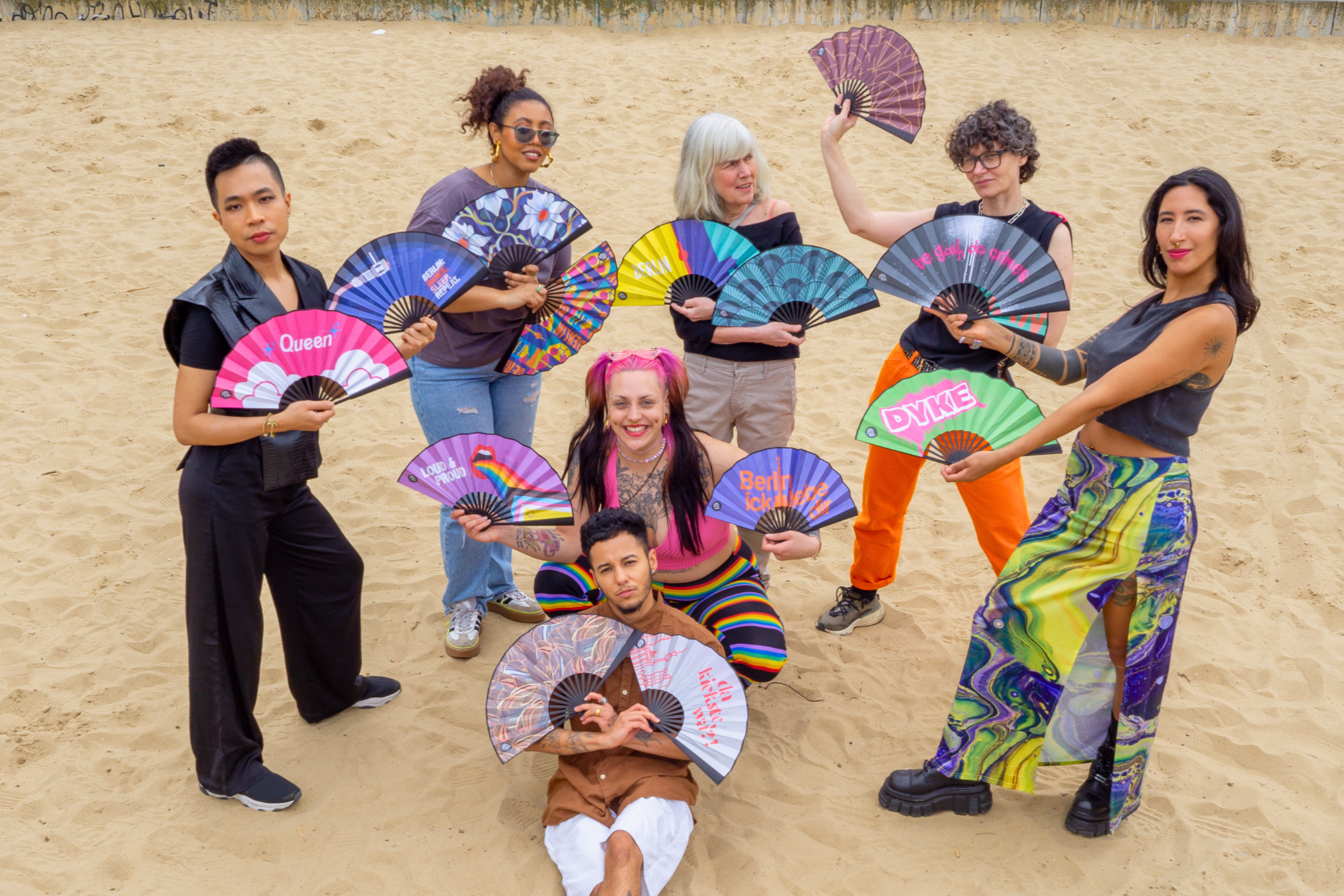 I'm a Fan! 15 Fächer mit Designs gehalten von sieben Personen sitzend bzw stehend im/ auf Sand.